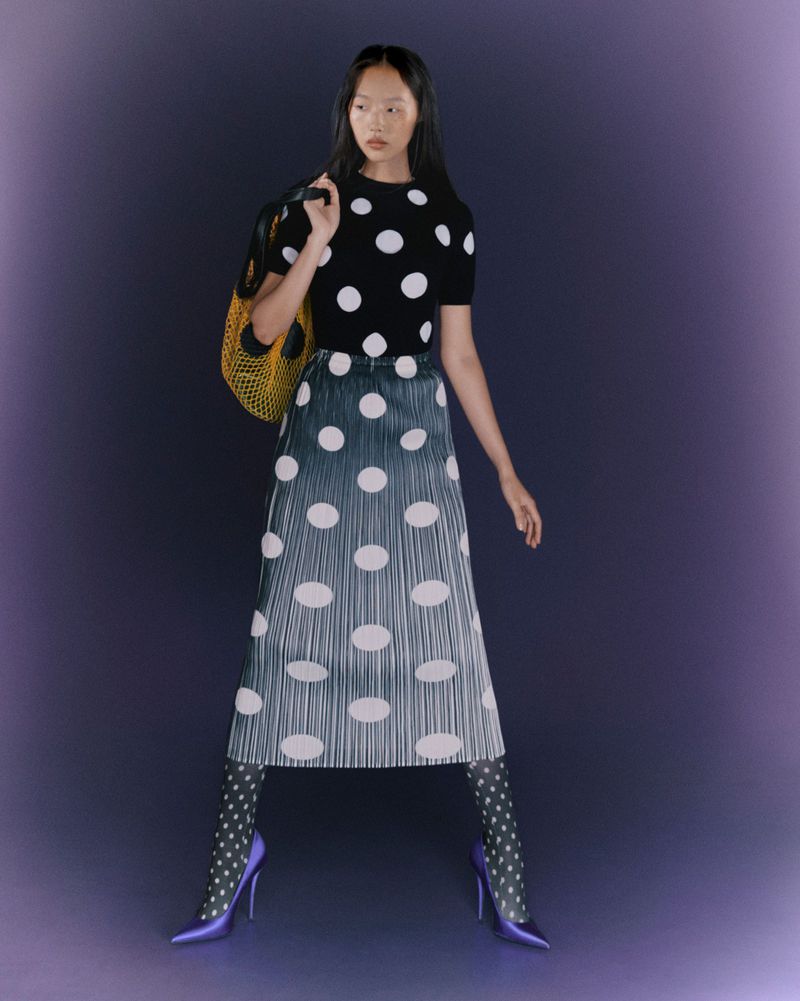 Young woman in polka-dot top, ombre skirt, and pointed heels with patterned tights poses in a studio against a purple backdrop.