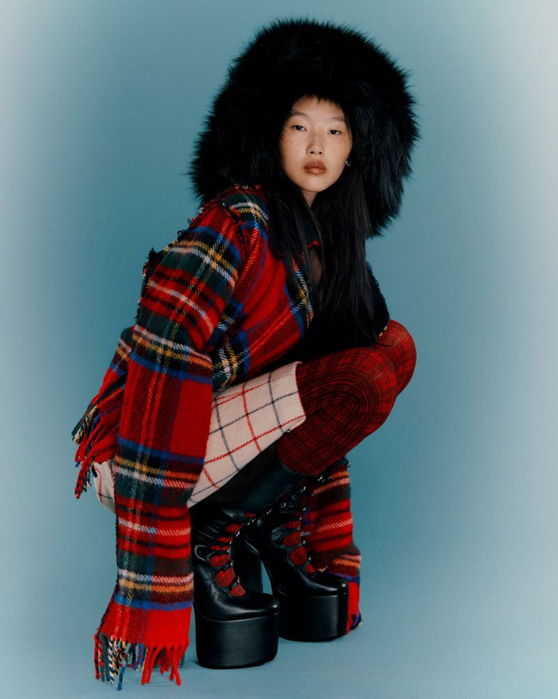 Asian woman in a squat pose wearing a large fur hat, plaid coat, and platform boots, set against a blue studio backdrop.