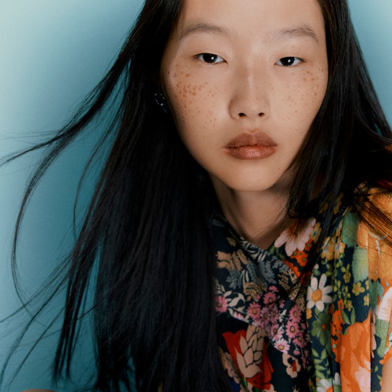 Close-up of an Asian woman with dark hair and freckles, wearing a floral print top against a light blue background in a studio shot.