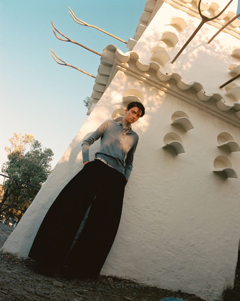 Stylish man in wide-leg pants leans against a white architectural structure outdoors with blue sky, trees, and dappled sunlight.