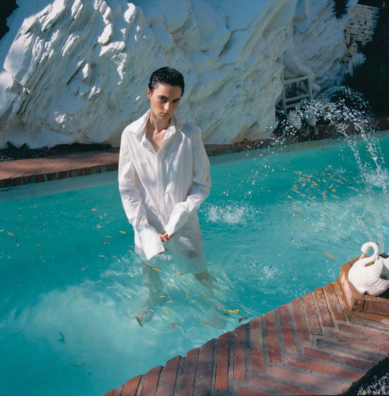 An adult stands in a turquoise pool wearing a white shirt, against a backdrop of a white, rocky structure and brick poolside border.