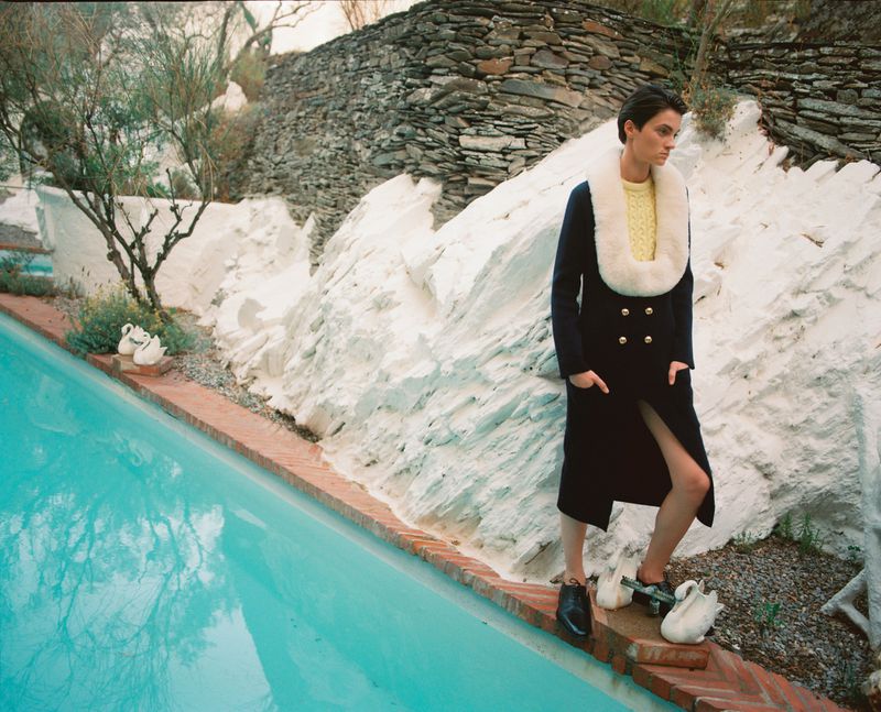 A woman in a dark coat and cream-colored sweater stands near a bright turquoise pool in a stylish outdoor setting with stone walls.