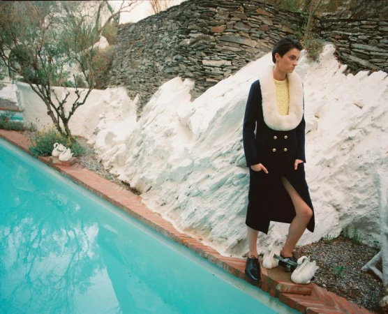 A woman in a dark coat and cream-colored sweater stands near a bright turquoise pool in a stylish outdoor setting with stone walls.