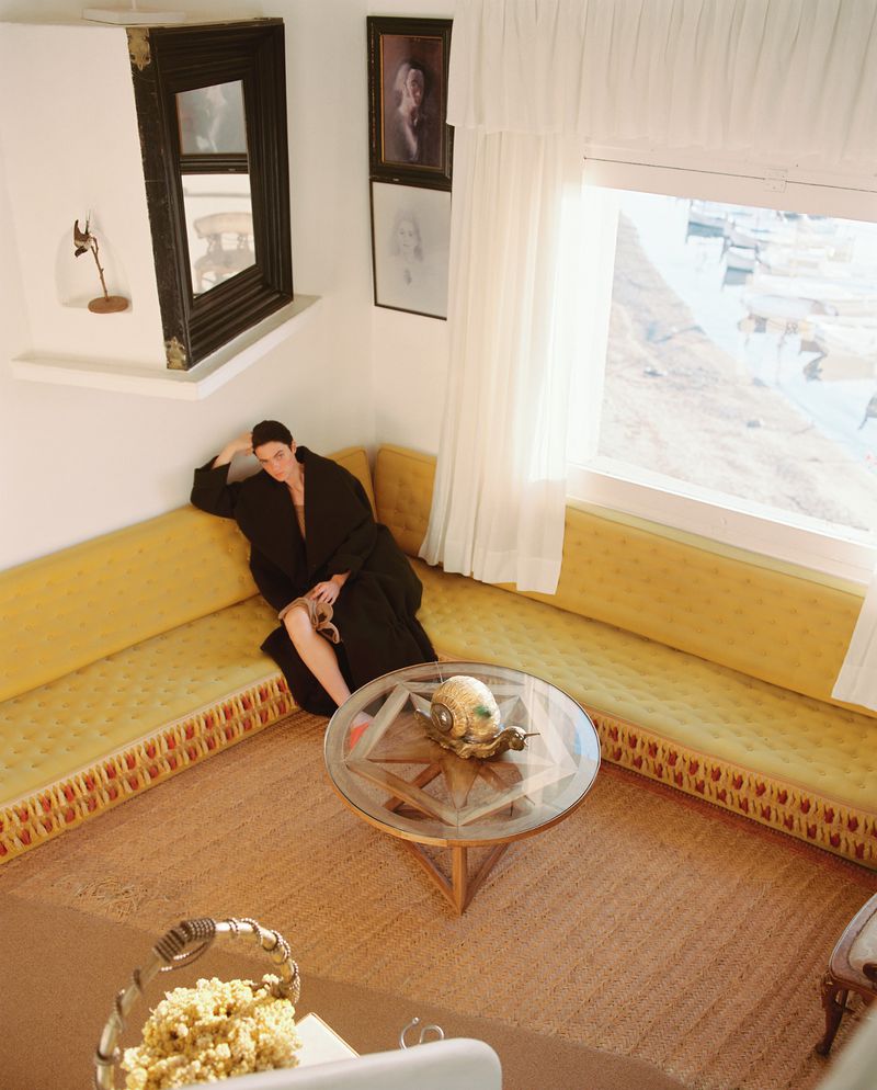 Indoor portrait of a woman in a black robe reclining on a yellow corner sofa in a room with a glass coffee table and white walls.