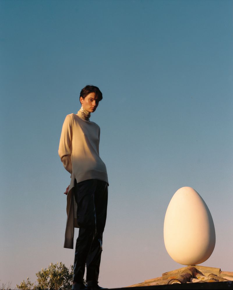 An adult stands next to a massive egg under a blue sky, bathed in soft light, creating a surreal and artistic fashion editorial.