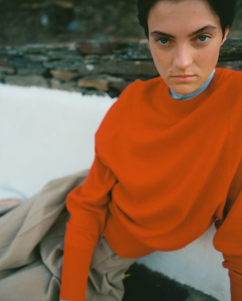 Close-up of a young adult woman with short dark hair and blue eyes, wearing an orange sweater, posing indoors with a serious expression.