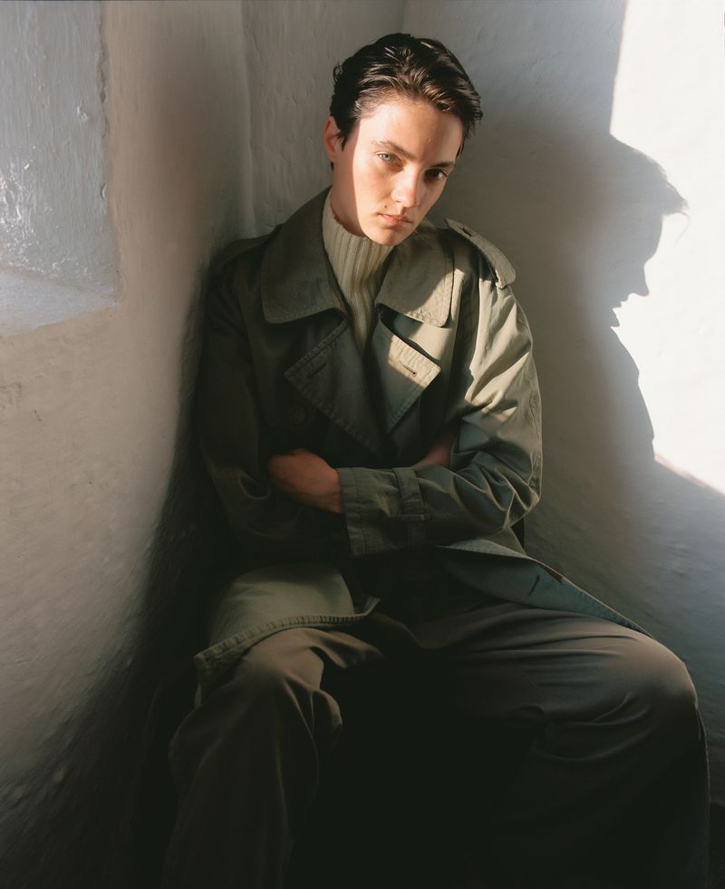 Young adult male in trench coat sitting in corner with natural sunlight and shadow.