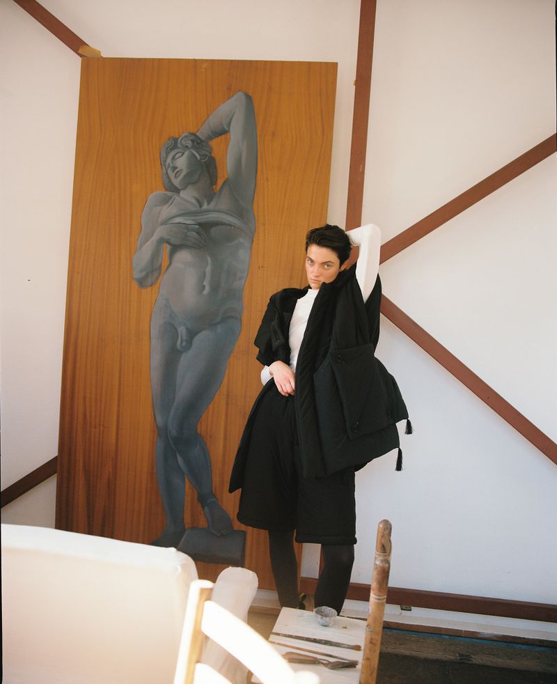 Person in avant-garde black fashion poses indoors by a large painting of a nude figure in a bright, high-contrast studio setting.