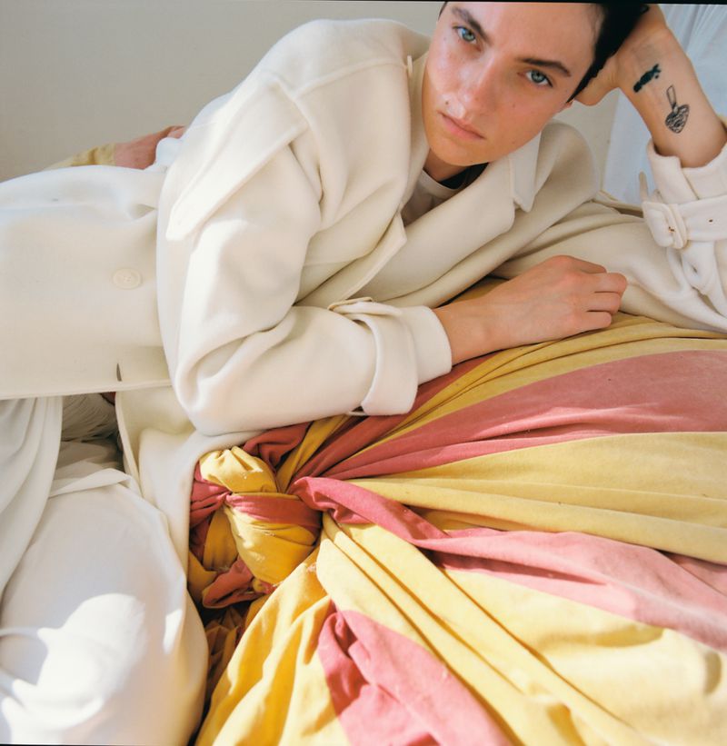 Elegant, young adult poses on a bed with colorful fabric accent. She wears a white coat, adding sophisticated touch to the portrait.