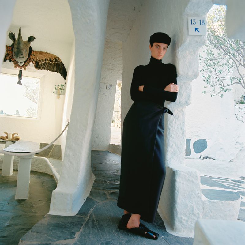 A woman in a black outfit stands in a white architectural setting with textured walls, an animal head sculpture, and stone flooring.