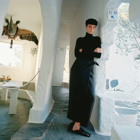 A woman in a black outfit stands in a white architectural setting with textured walls, an animal head sculpture, and stone flooring.