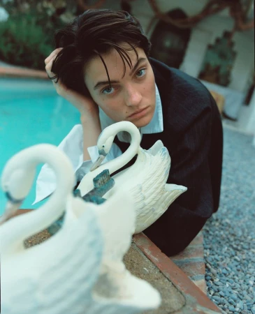 Eye-level portrait of a young adult woman in a blazer, resting her head on her hand, posing near swan statues and a blue pool outdoors.