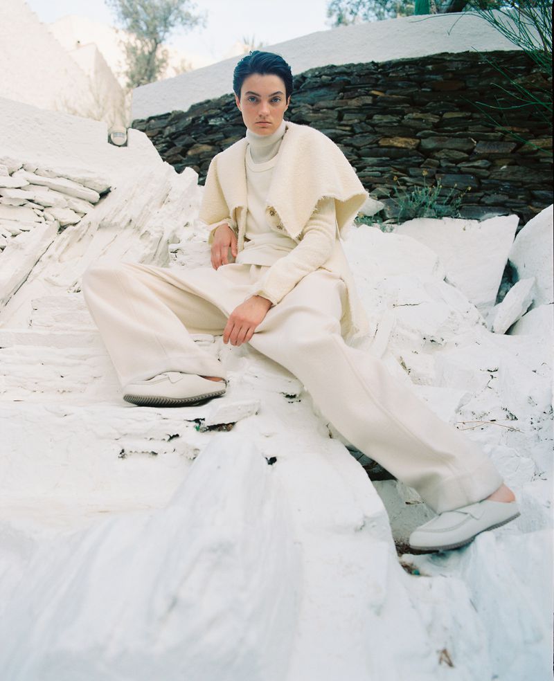 Woman in neutral monochrome fashion poses on white rocks outdoors, wearing a cape, turtleneck, pants, and loafers in full shot.