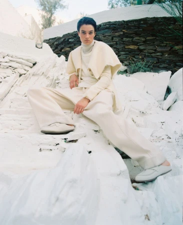 Woman in neutral monochrome fashion poses on white rocks outdoors, wearing a cape, turtleneck, pants, and loafers in full shot.