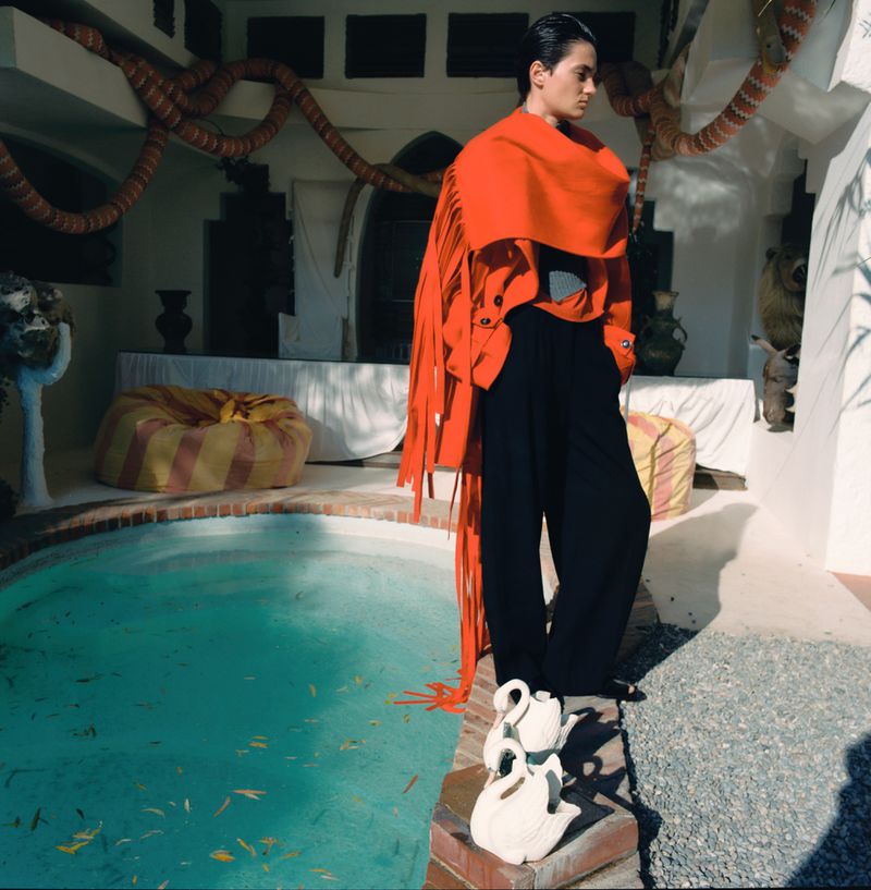 Fashion shot of an adult male in black pants and an orange shawl standing next to a pool with white swan objects in a courtyard.