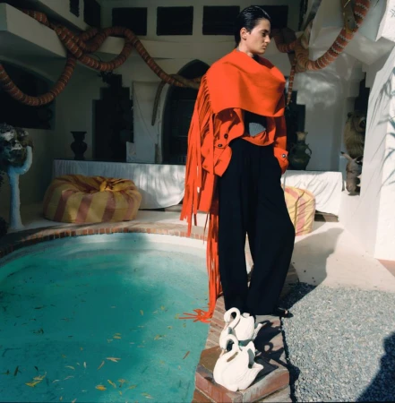 Fashion shot of an adult male in black pants and an orange shawl standing next to a pool with white swan objects in a courtyard.