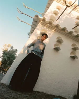 Model standing outdoors leaning against a white stucco building wearing a light blue top and dark trousers.