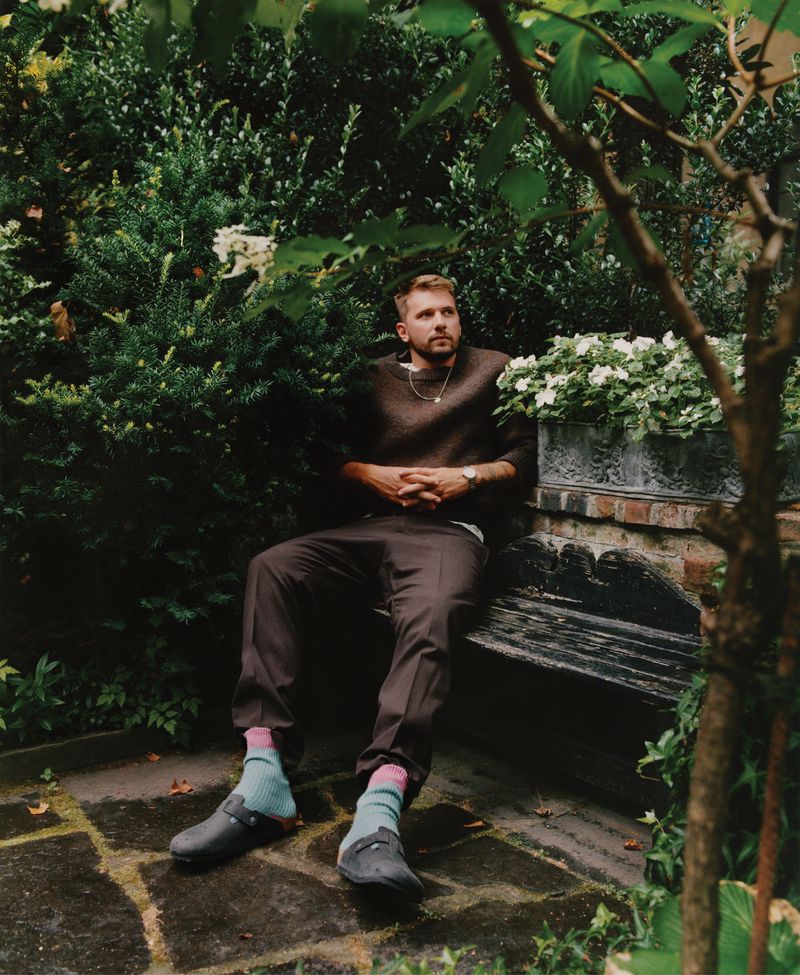 Blond man with a beard poses while sitting on a bench in a lush outdoor garden setting with greenery and textured stone ground.