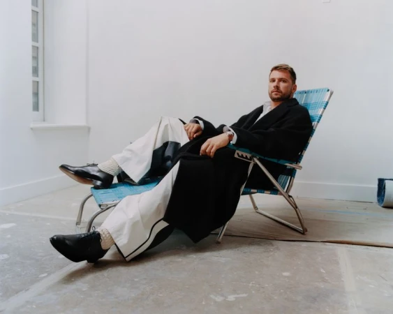 Adult male model with a beard is relaxing in a folding chair while wearing a black coat in a minimalist photo studio setting.
