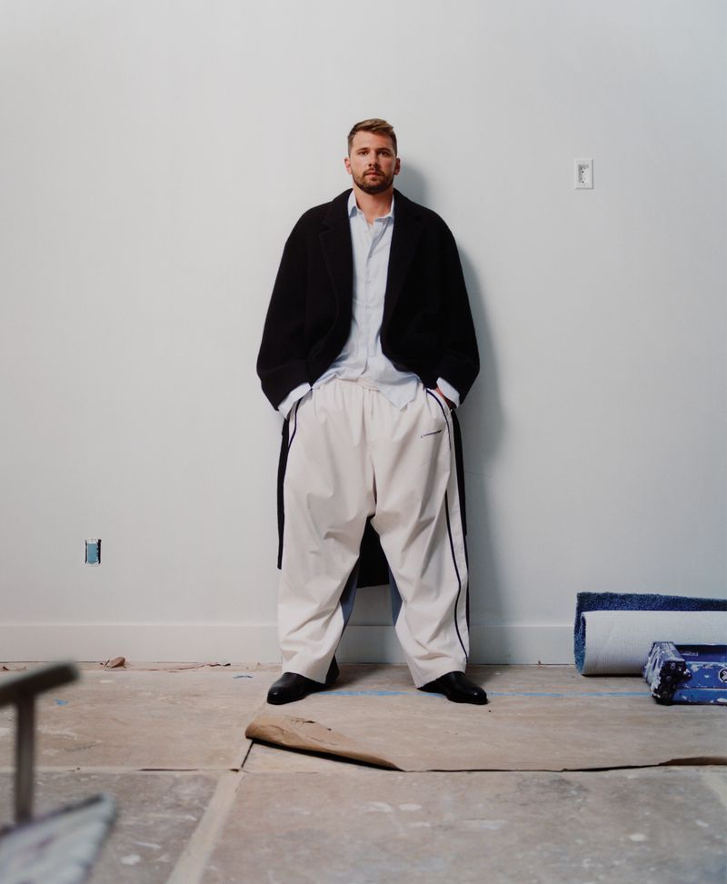 A man poses in a studio setting, dressed in a light shirt, white pants, and a dark coat. He has his hands in his pockets.