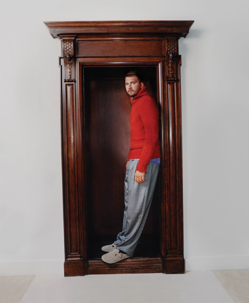 Adult man in a red sweater and grey pants stands inside an ornate, dark-wood doorframe against a light grey wall in a studio shot.