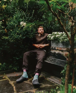 Man seated on a garden bench wearing a brown sweater, trousers, pale blue socks, and clogs surrounded by lush greenery.