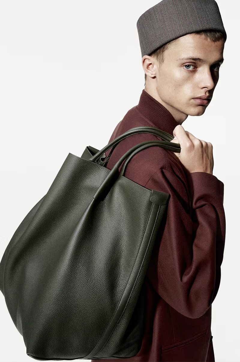 Fashion shot of a slender adult male in burgundy top and grey hat, carrying a large green leather tote bag over his shoulder.