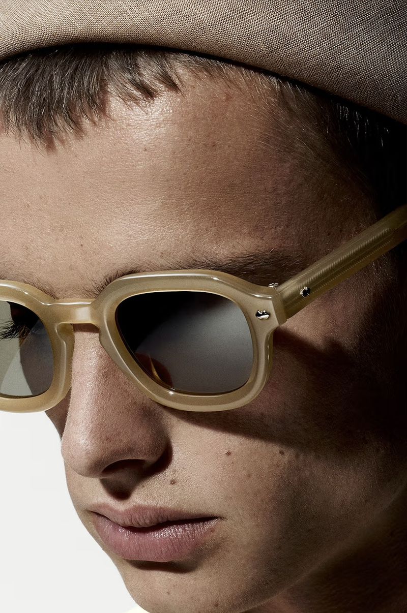 Close-up of a person wearing sunglasses and a hat, focusing on the details of the glasses and facial features in a neutral setting.