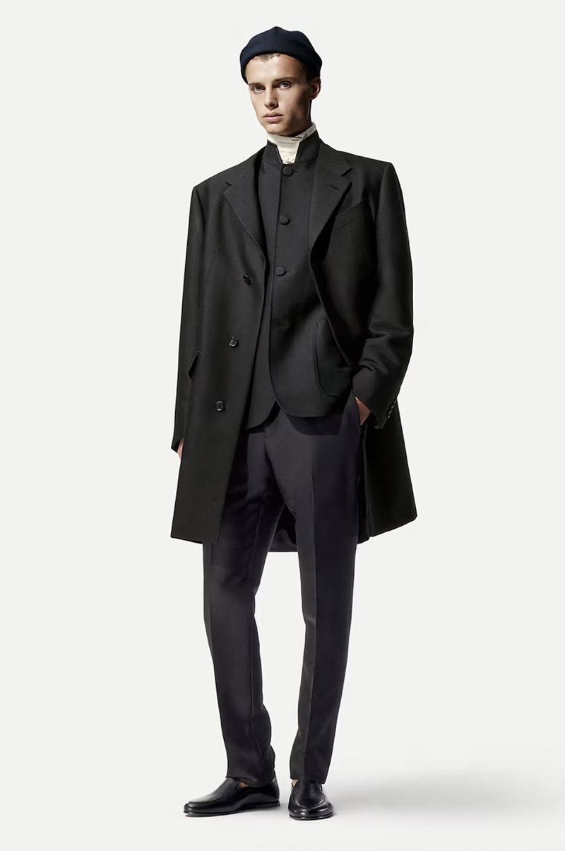Male fashion model in a black overcoat, vest, trousers, turtleneck, and beanie stands against a white backdrop in a full shot.