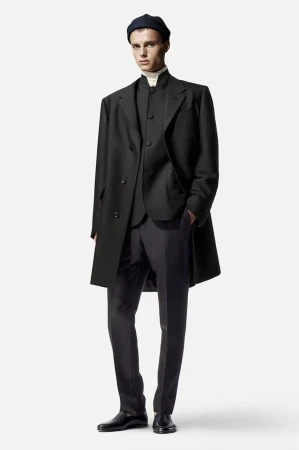 Male fashion model in a black overcoat, vest, trousers, turtleneck, and beanie stands against a white backdrop in a full shot.