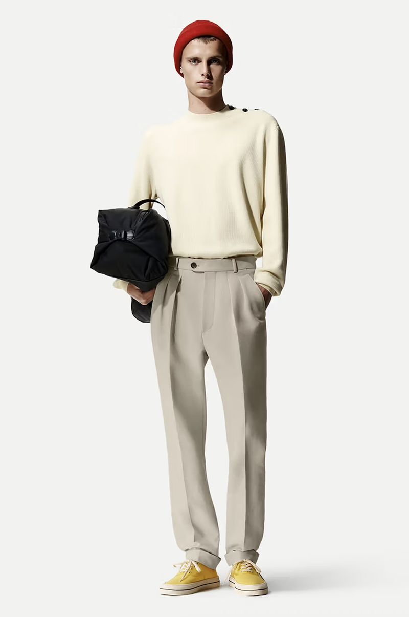 Full-shot of a young fair-skinned man wearing a cream sweater, beige pants, yellow sneakers, and red beanie, holding a black backpack.