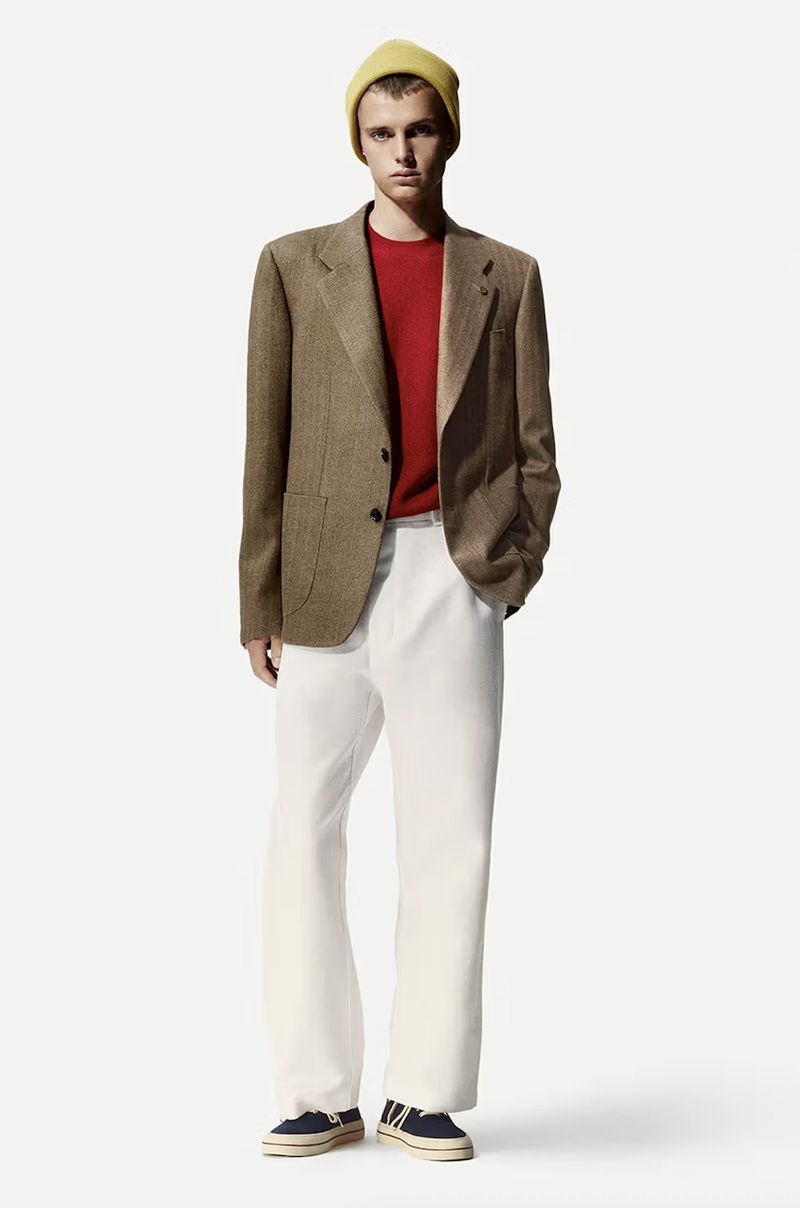 Fashion portrait of a young man wearing a tan blazer, red sweater, white pants, yellow beanie, and sneakers on a white background.