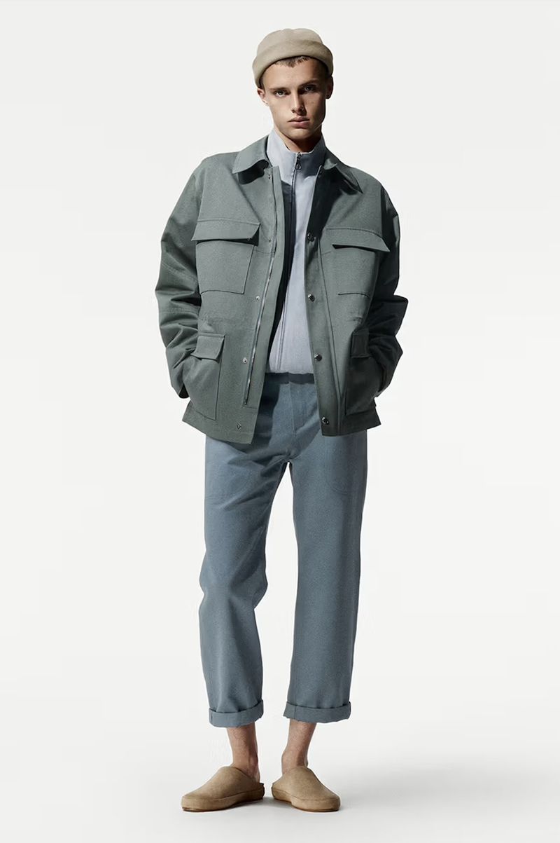Male fashion model poses in a studio wearing a green jacket, light blue shirt and pants, paired with tan-colored slippers and a beanie.