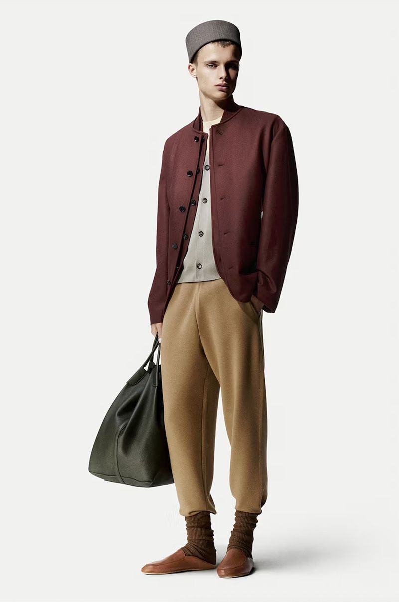 Male fashion model wears burgundy jacket, beige trousers, brown socks and shoes, holding an olive green leather bag in a studio shot.