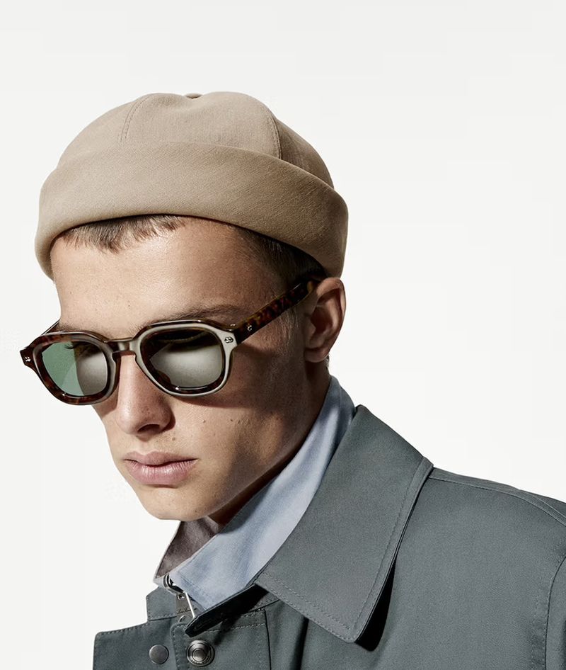 A stylish young man poses in a studio setting, wearing sunglasses, a brown hat, and a gray jacket, against a clean white backdrop.