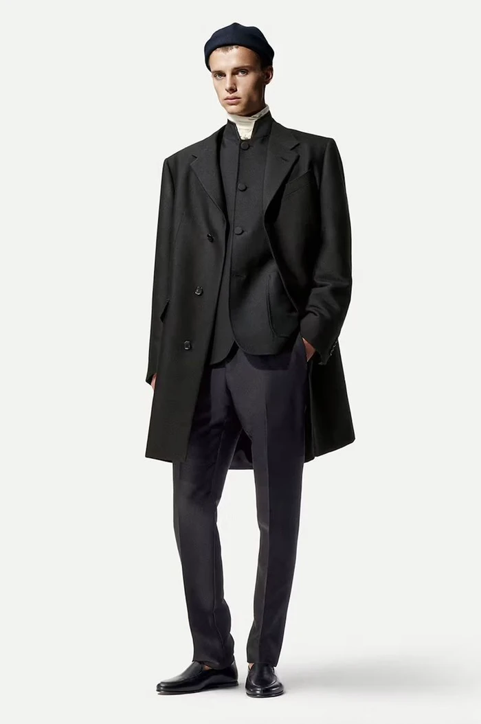 Model standing against a white seamless background wearing a dark coat, dark suit jacket, trousers, and a beanie hat