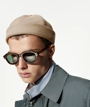 Model wearing tortoiseshell sunglasses, beige cap, and slate grey coat against a bright white background