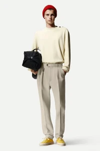 Model wearing a red beanie, cream sweater, light trousers and yellow sneakers while holding a black bag