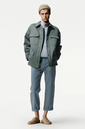 Model wearing a grey jacket, blue trousers, and beige beanie standing against a white seamless background.
