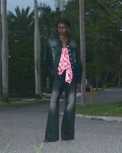 A woman in a denim jacket and flared jeans poses on a street lined with palm trees under a twilight sky.