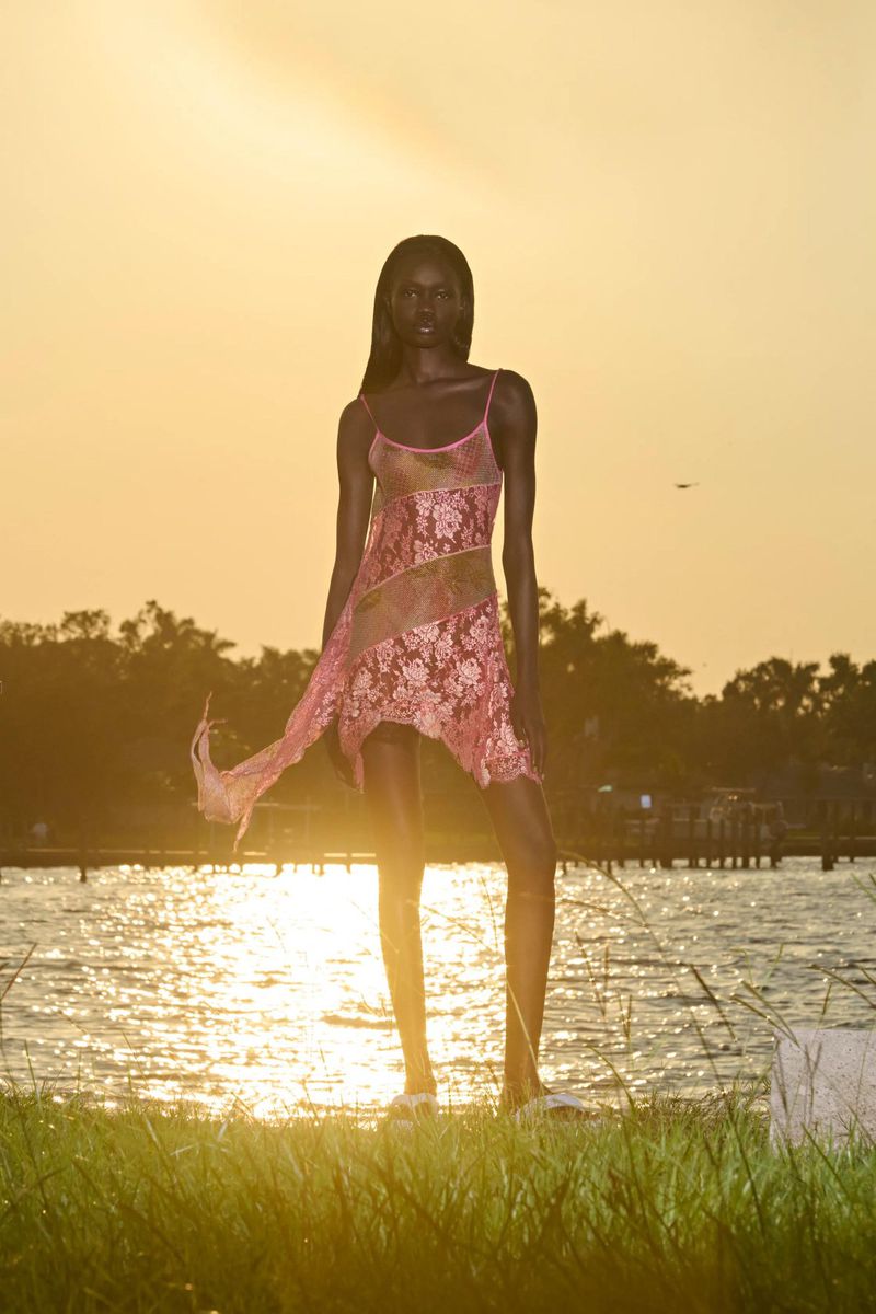 Woman in pink floral dress poses near grassy water's edge against warm golden hour light, creating a dreamy, tranquil fashion portrait.