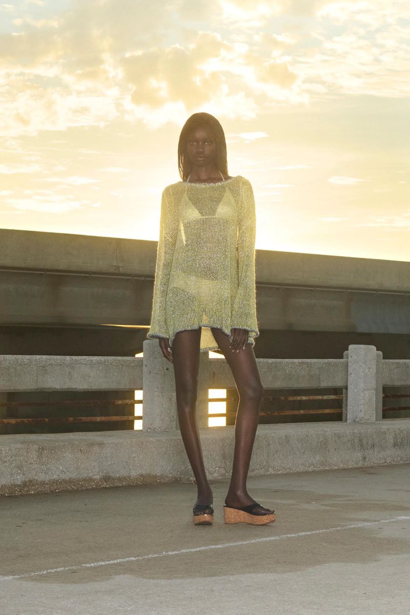 A slender dark-skinned woman poses outdoors in a sheer, long-sleeved cover-up and sandals against a background of concrete and sunset sky.