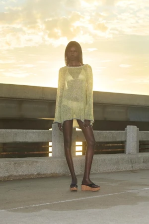 A slender dark-skinned woman poses outdoors in a sheer, long-sleeved cover-up and sandals against a background of concrete and sunset sky.