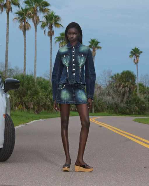 Fashion model in denim jacket and skirt poses on road near car, backed by palm trees and sky for outdoor fashion editorial shoot.