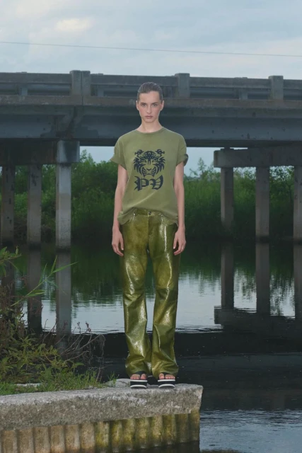 A woman stands on a concrete structure by a waterway wearing an olive t-shirt, shiny olive pants, and sandals under an overcast sky.