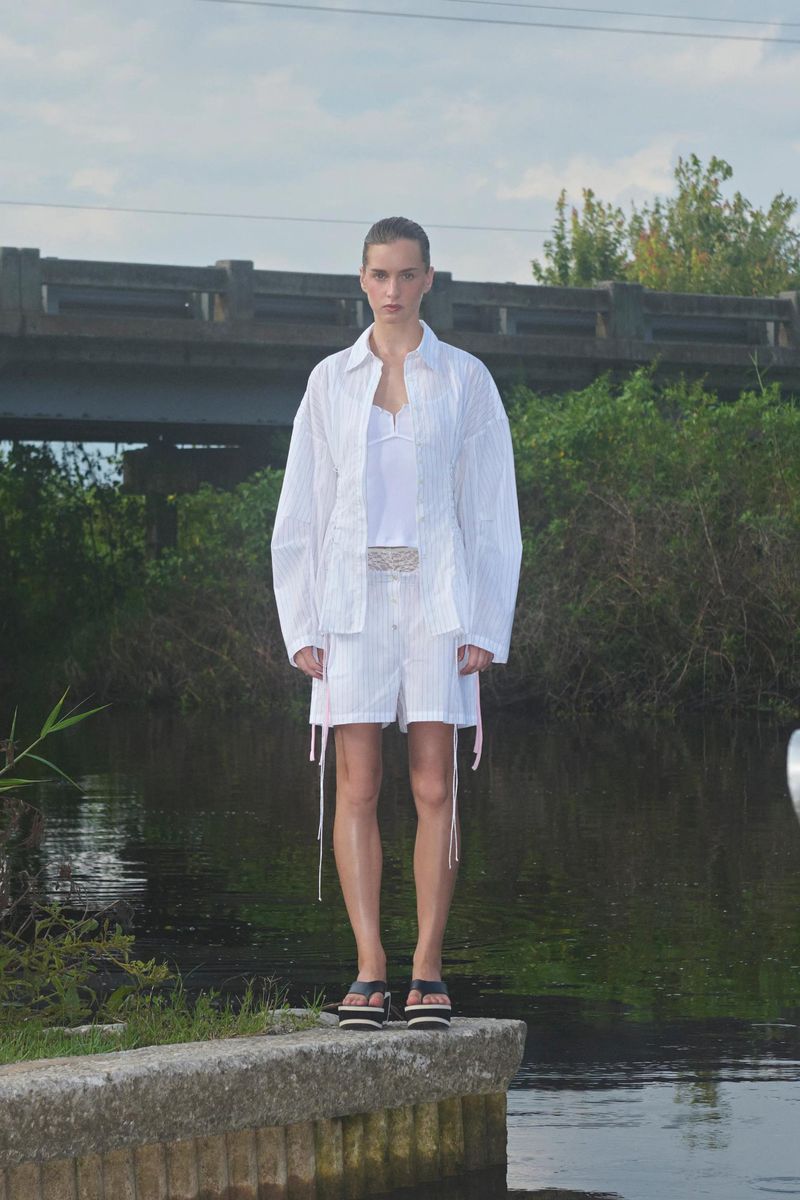 Fashion model in white striped shorts suit stands on a concrete ledge by a river beneath a bridge in natural light.