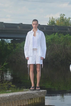 Fashion model in white striped shorts suit stands on a concrete ledge by a river beneath a bridge in natural light.