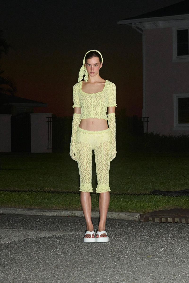Woman in crocheted yellow top, capri pants, gloves, and white platform sandals poses in front of a pink house at dusk.