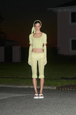 Woman in crocheted yellow top, capri pants, gloves, and white platform sandals poses in front of a pink house at dusk.