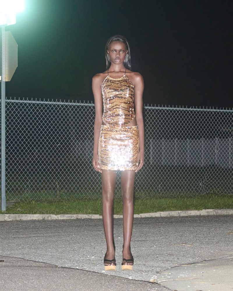 Elegant adult woman in a shimmering halter dress and heels poses in an outdoor nighttime fashion shot against a chain-link fence.